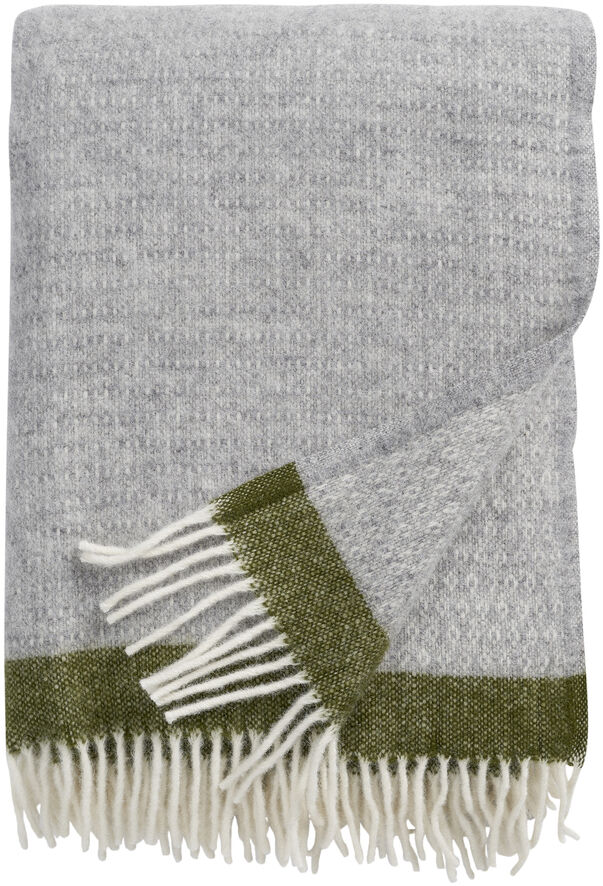 Ein großes, rechteckiges Plaid Hampus in Grau mit weißen und grünen Fransen liegt zusammengefaltet auf einem weißen Hintergrund. Das robuste Strickmuster und die gedeckten Farben passen gut in ein gemütliches Zuhause.