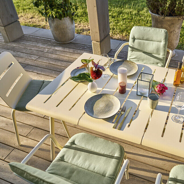 Ein moderner Esstisch für den Außenbereich, »VAPIO« 180x90 cm, mit grünen Kissen und kompletter Tischdekoration, arrangiert auf einer Terrasse mit Blick auf einen grünen Garten. Aus Holzplatten und Metallstühlen mit grünen Polstern gefertigt.