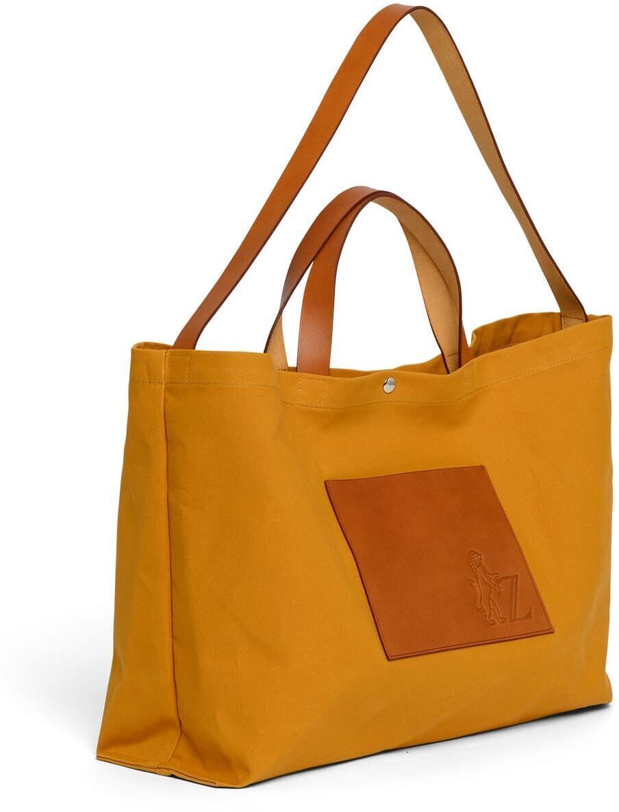 Die Jubiläums-Markttasche ist eine große, robuste Stofftasche in gelber Farbe mit braunen Ledergriffen und einer braunen Vordertasche. Ideal für den Einkauf auf dem Markt oder als stilvolles Accessoire.