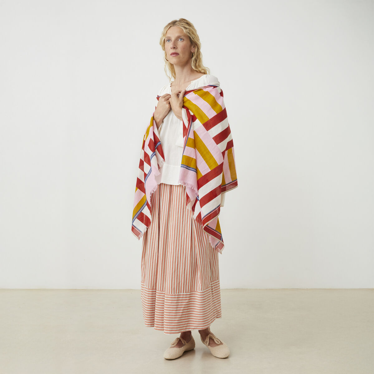 Eine Frau trägt einen gestreiften Baumwollschal »No. 771« von MOISMONT in Rot, Weiß, Gelb und Rosa. Dazu kombiniert sie einen weiß-roten Rock und ein weißes Oberteil. Sie steht barfuß auf hellem Boden vor weißer Wand. Das Bild wirkt hell und freundlich.