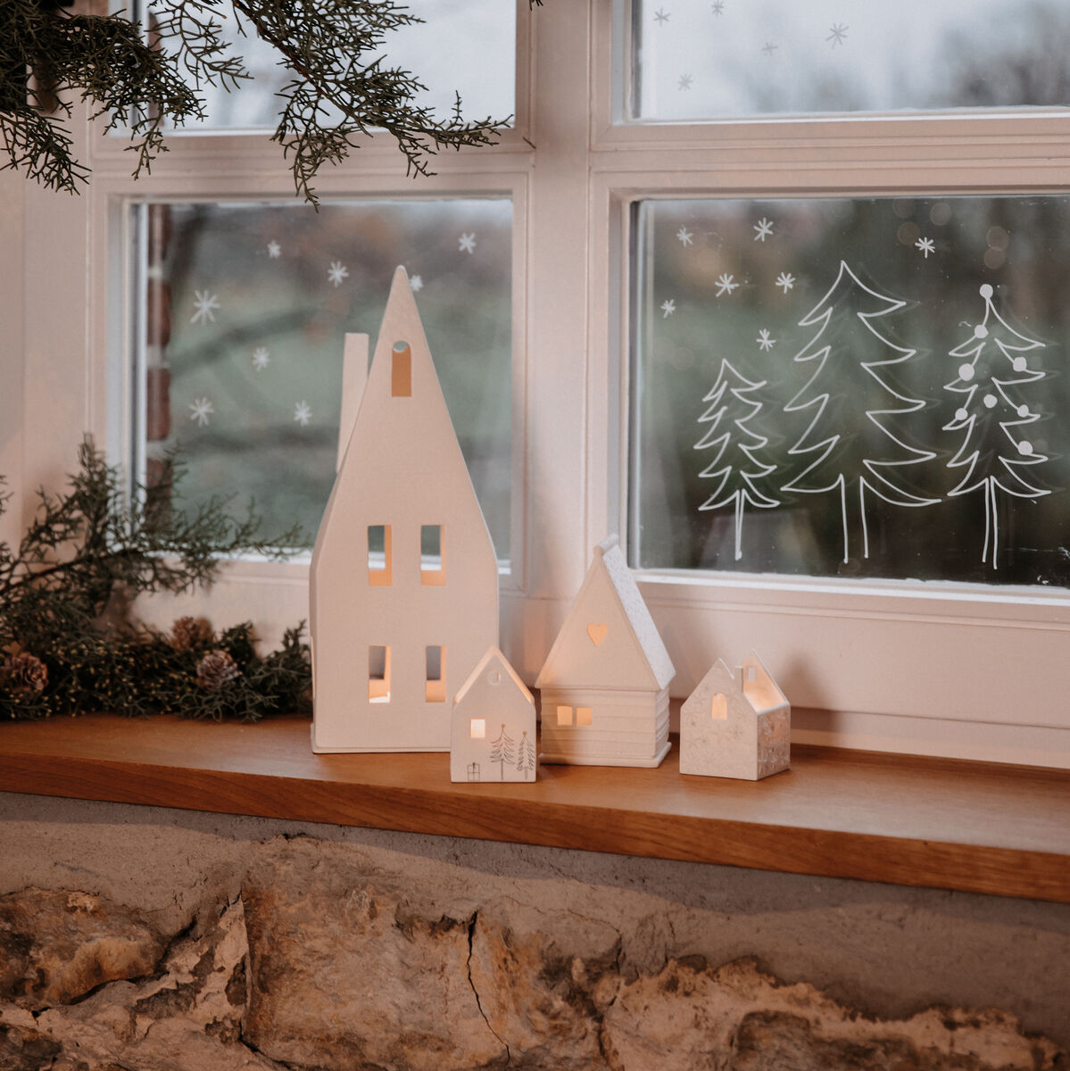Am Fensterbrett steht ein großes Lichthaus mit schmalen, hohen Fenstern, das von kleineren Häusern aus Keramik flankiert wird. Im Hintergrund sind Tannen- und Schneeflocken-Dekorationen auf die Fensterscheiben gemalt. Ein grüner Tannenzweig ist oben am Fensterrahmen drapiert.