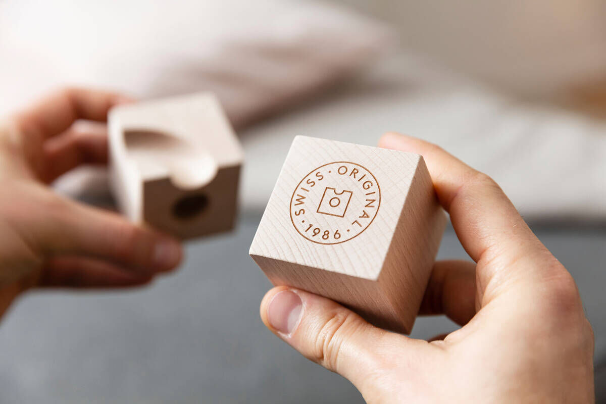 Nahaufnahme zweier Hände, die zwei Holzbausteine mit eingraviertem 'Swiss Original 1986'-Stempel halten. Die Bausteine sind aus hellem Holz gefertigt und haben eine glatte Oberfläche.