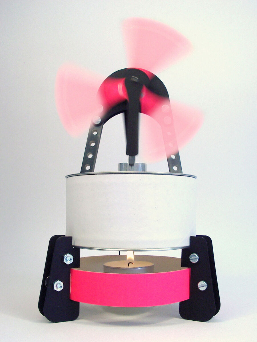 Ein Bausatz für einen Stirling Motor, der mit einem Teelicht angetrieben wird. Der Motor verfügt über schwarze und silberne Bauteile mit einem pinken Akzentband in der Mitte. Die Flügel des Motors scheinen in einer roten Bewegung dargestellt zu sein. Der Hintergrund ist weiß.
