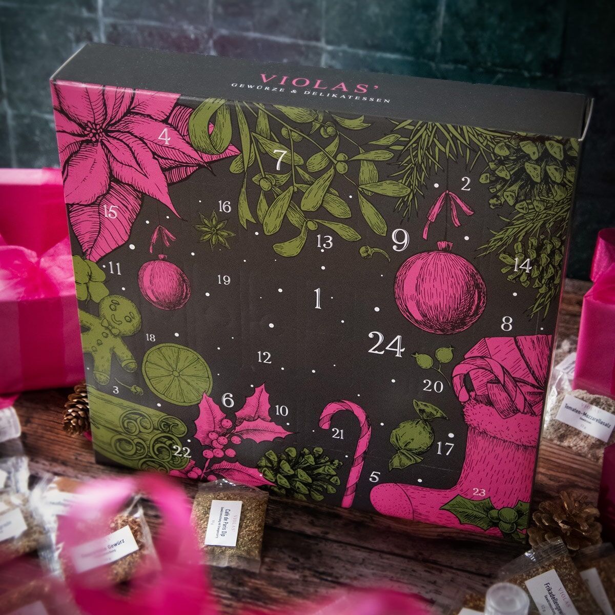 Violas Adventskalender mit schwarzem Hintergrund, pinken Poinsettien und grünen Mistelzweigen. Die Box zeigt Zahlen für jeden Tag des Advents und ist umgeben von weiteren weihnachtlichen Dekorationen und Geschenken in Pink.