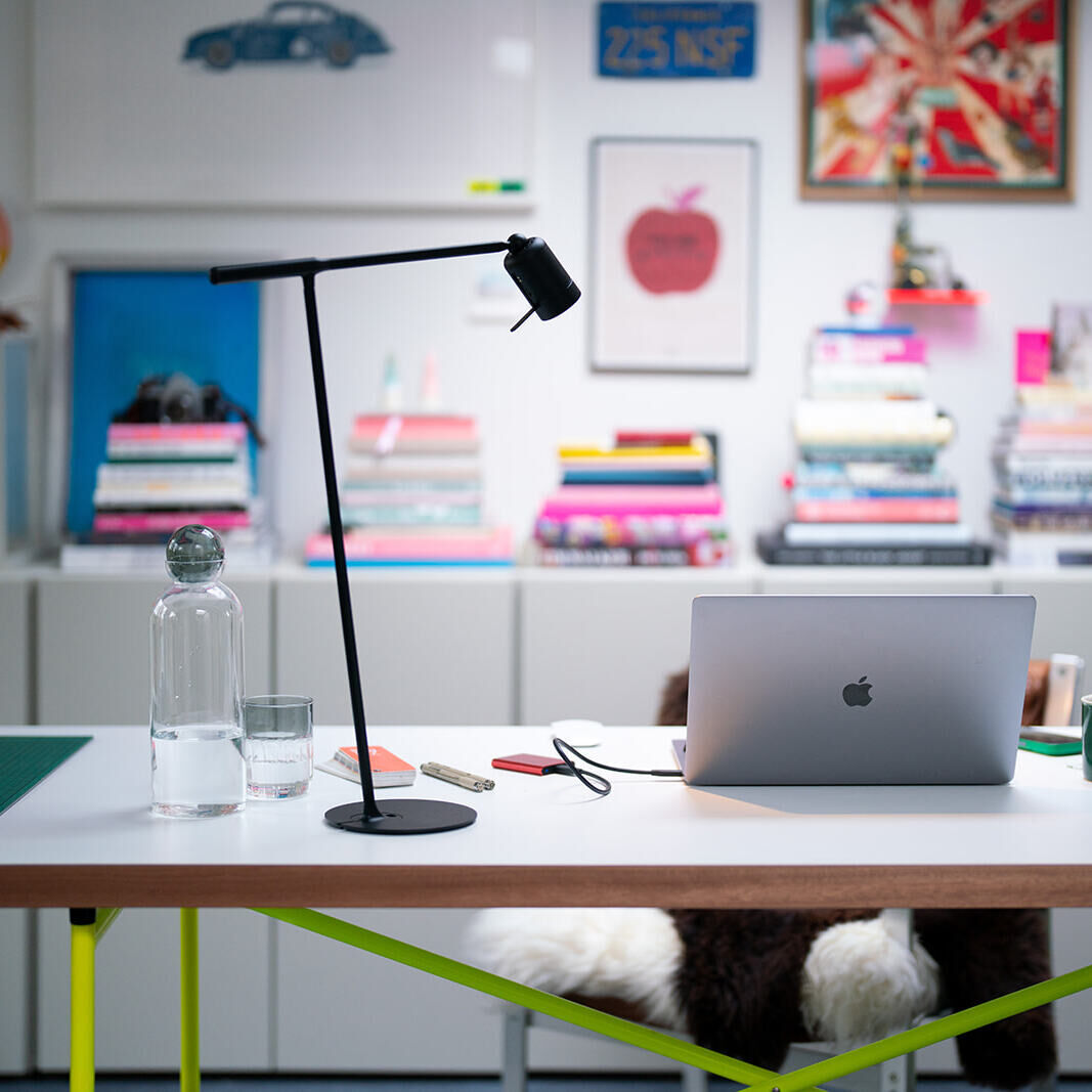 Ein moderner Schreibtisch mit einer schlanken schwarzen Schreibtischlampe 'Leuchte »ONE« mit Halterung'. Auf dem Tisch stehen ein Laptop, ein Glas Wasser, eine Kanne, einige Büroartikel und Bücher. Im Hintergrund sind bunte Bücherstapel und mehrere gerahmte Bilder und Poster an einer weißen Wand zu sehen.