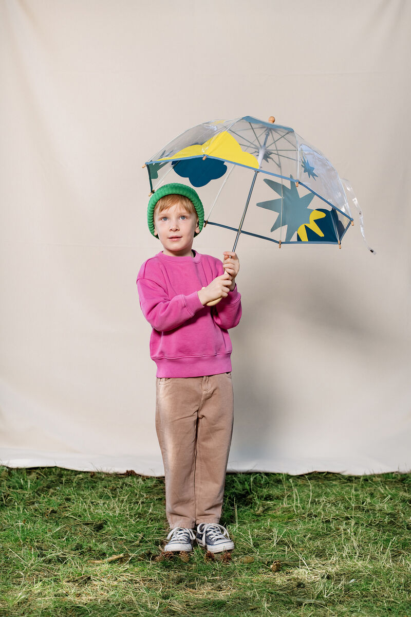 Ein Junge trägt einen transparenten Regenschirm mit blauer und gelber Wolken-Dekoration. Der Junge trägt einen grünen Hut, einen rosa Pullover und beige Hosen. Er steht auf einer Wiese vor einem beigen Hintergrund.