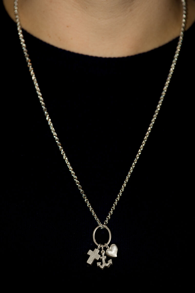 Eine silberne Kette mit drei Anhängern: einem Kreuz, einem Anker und einem Herz. Die Kette wird um den Hals einer Person getragen, die einen schwarzen Hintergrund hat.