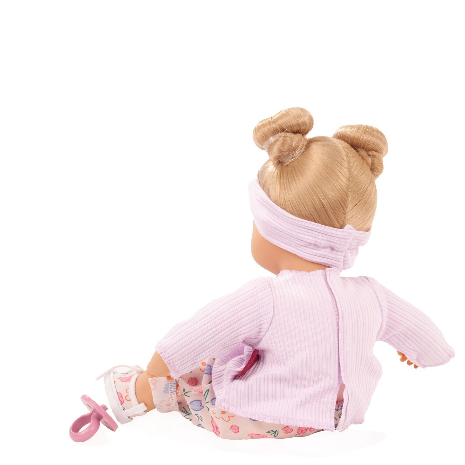 Die Abbildung zeigt eine Babypuppe mit zwei Zöpfen, die ein lilafarbenes Oberteil, gemusterte Hose und ein Stirnband trägt. Die Puppe sitzt auf dem Boden und blickt nach hinten. Rechts am Arm ist ein Schnuller befestigt.