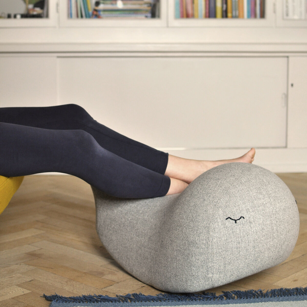 Ein grauer Schaukelwal, der als Kindermöbelstück dient, in einem Wohnzimmer mit Holzboden und Bücheregal im Hintergrund. Ein Kind liegt mit den Beinen auf dem Spielzeug, was Bequemlichkeit und Verspieltheit veranschaulicht. Das Bild zeigt eine gemütliche und moderne Inneneinrichtung.
