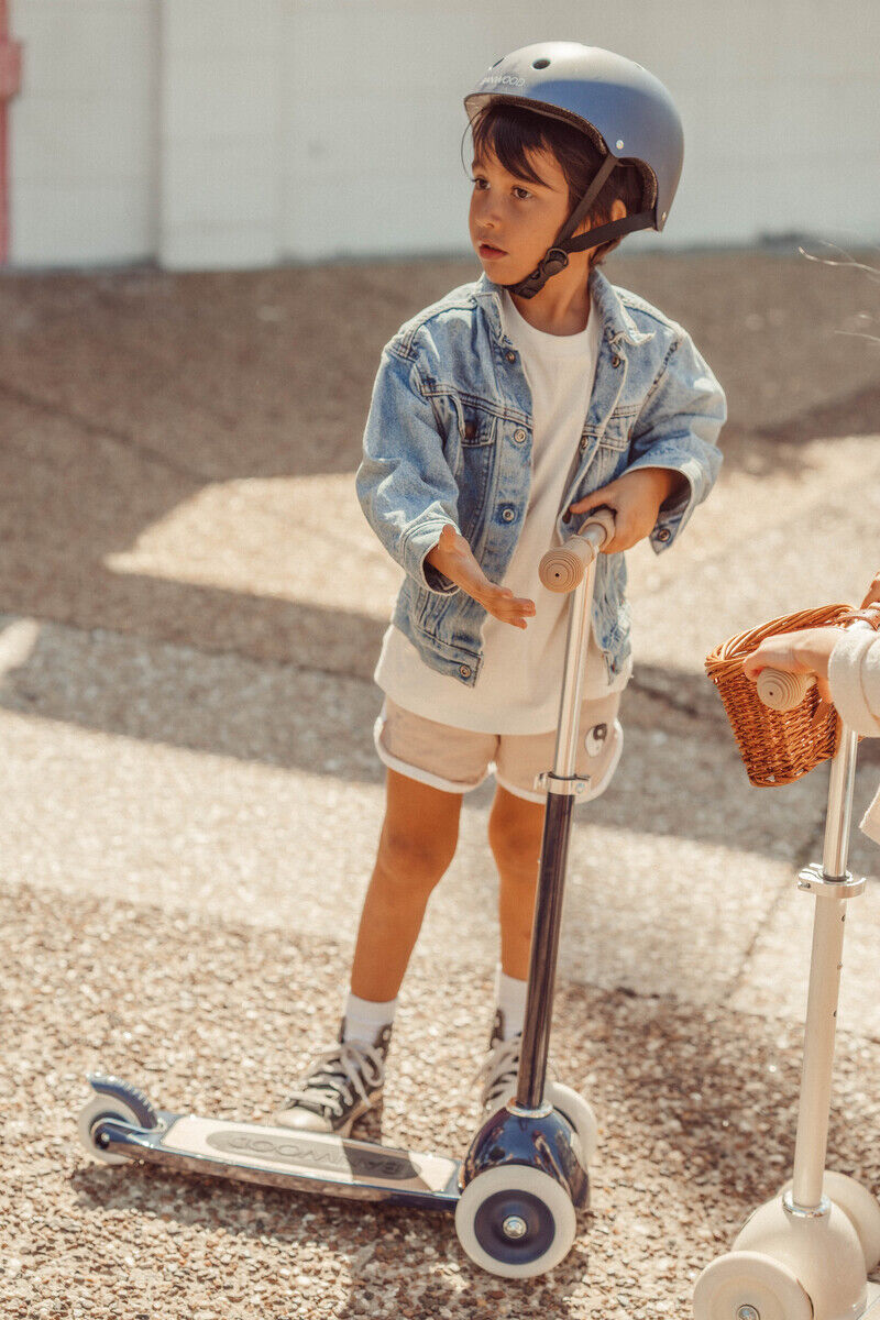Ein kleiner Junge mit Helm und blauer Jeansjacke fährt einen blauen Kinder Scooter. Er trägt weiße Shorts und schwarze Schuhe. Im Hintergrund ist ein anderer Scooter zu sehen.