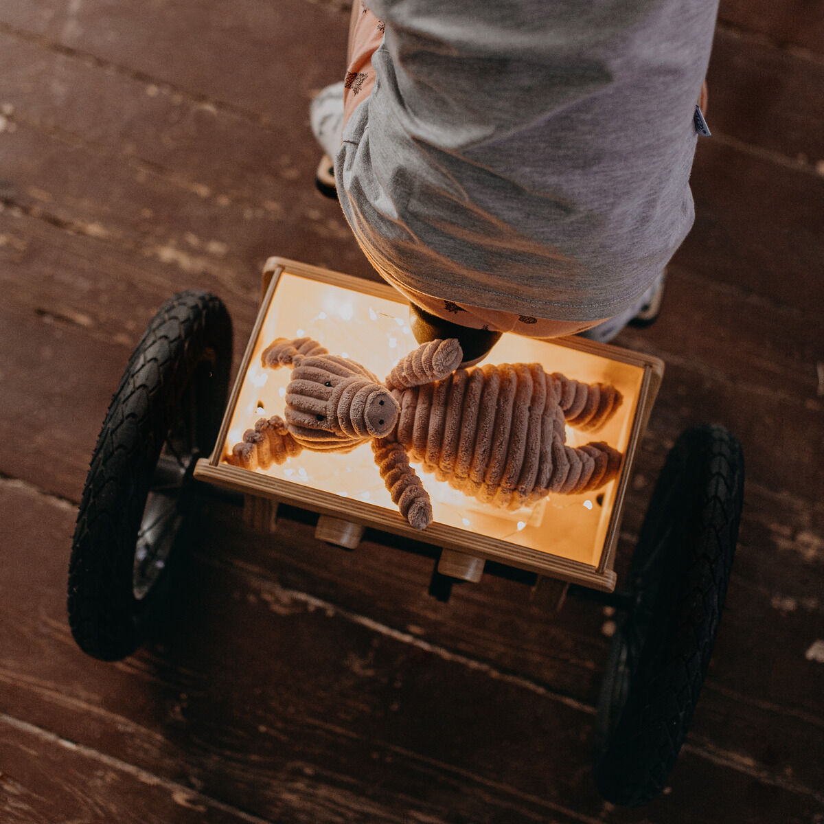 Ein Kind sitzt auf einer Erweiterung zum Dreirad für ein Balance Bike. Das Kind fährt auf einem holzfarbenen Boden, und auf einer Spiegelplatte vor sich liegt ein Stofftier. Die Aufnahme zeigt das Produkt im Alltagsgebrauch in einer Innenraumszene.