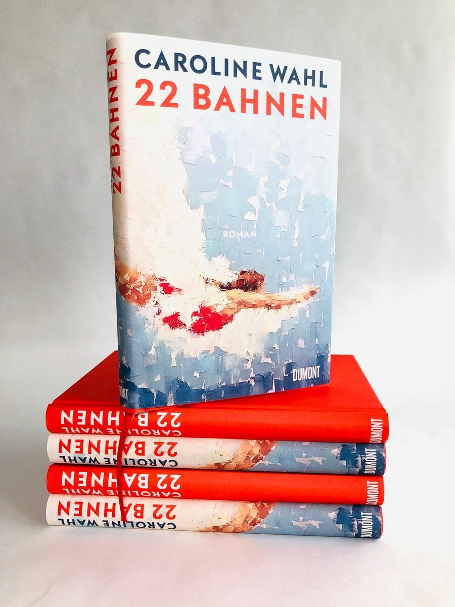 Buchcover von '22 Bahnen' von Caroline Wahl ist auf einem Stapel von vier weiteren Büchern desselben Titels zu sehen. Das Cover ist in Blau- und Weißtönen gehalten und zeigt ein abstraktes Kunstwerk einer schwimmenden Person. Der Titel '22 Bahnen' und der Autorenname 'Caroline Wahl' sind in roten und schwarzen Buchstaben abgebildet.