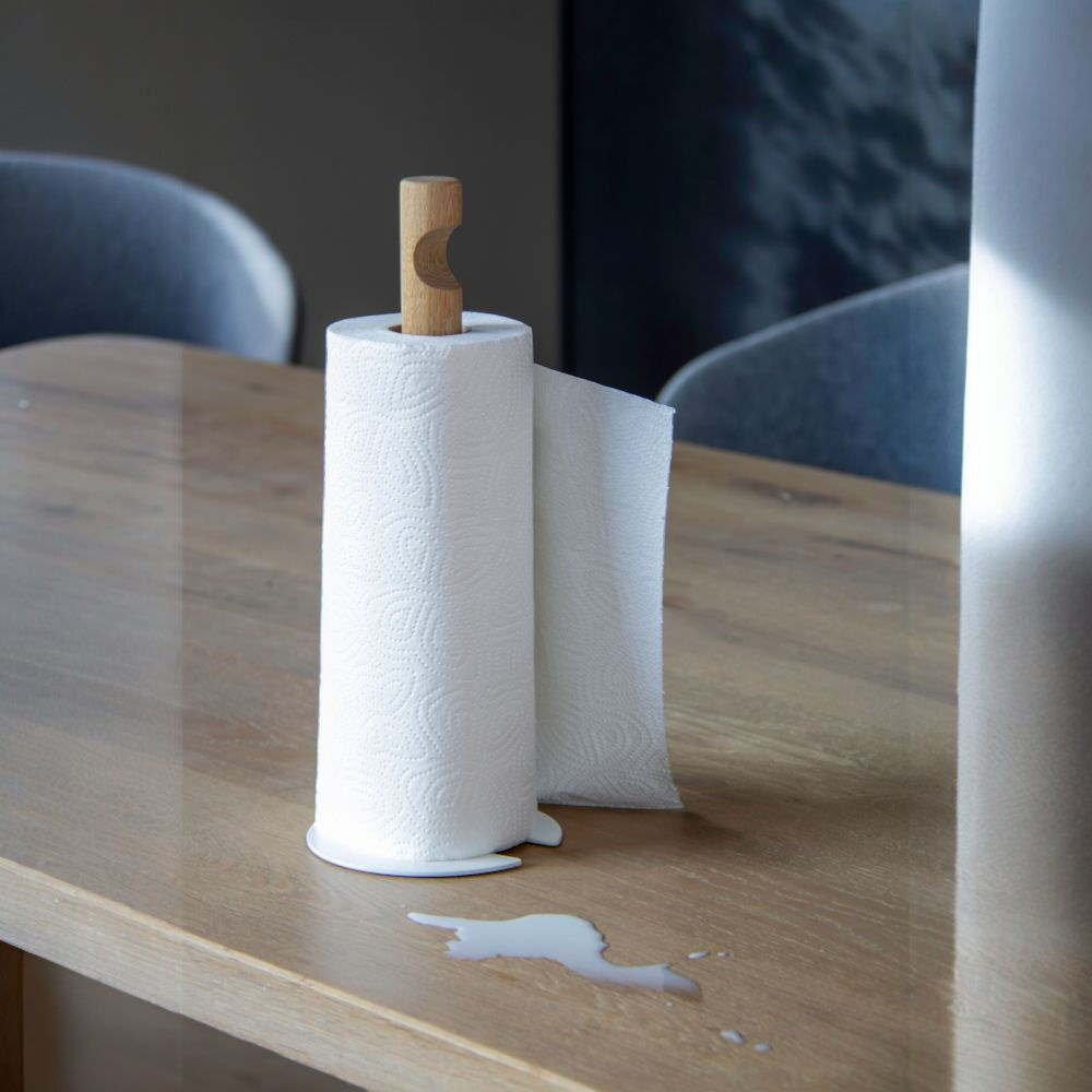 Küchenrollenhalter GIVE von finbeaux auf einem hellen Holztisch: Eine weiße, geprägt gemusterte Küchenpapierrolle steckt auf einem senkrechten Stab aus hellem Holz mit geschwungenem Griff. Ein Blatt hängt seitlich herab; davor liegt eine kleine verschüttete weiße Flüssigkeit. Im Hintergrund stehen graublaue Polsterstühle.