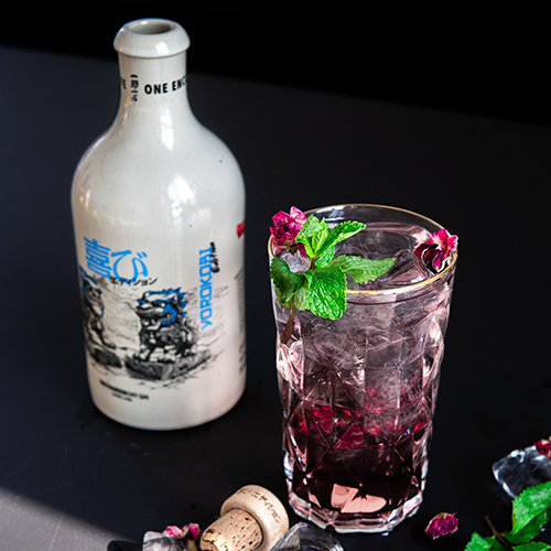 Glas mit lila Cocktail, Eiswürfeln, Minzblättern und kleinen Blüten, daneben eine weiße Flasche mit japanischer Schrift und Korken