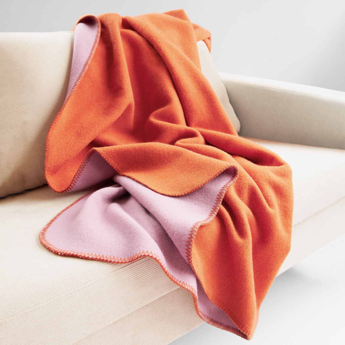 Eine hochwertige Merinowolldecke »DUO« von Eagle Products liegt dekorativ auf einem hellen Sofa. Die Decke zeigt zwei Seiten: Eine kräftig orangefarbene und eine fliederfarbene. Das Material wirkt weich und voluminös, ideal für gemütliche Wohnräume oder als stilvolles Geschenk.