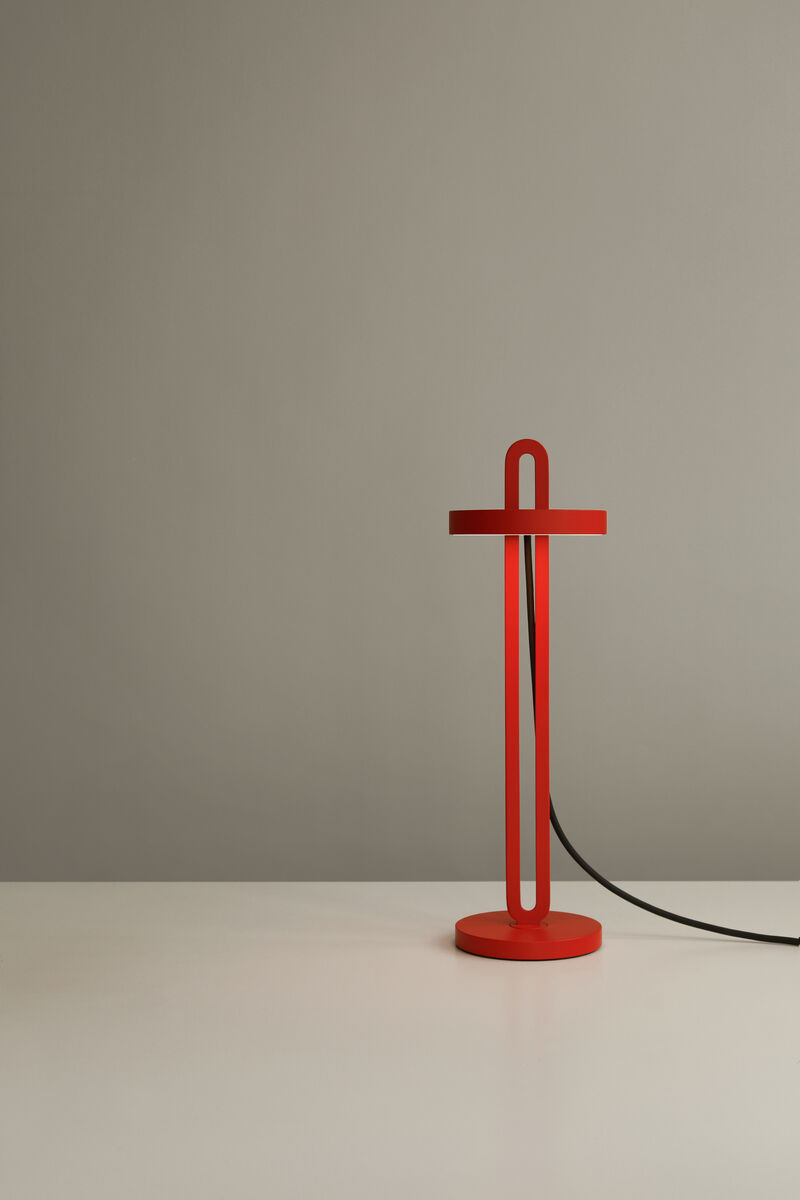Eine rote Tischlampe der Marke Echo steht auf einem weißen Tisch vor einer grauen Wand. Die Lampe hat ein schlichtes, modernes Design mit einem schwarzen Kabel.