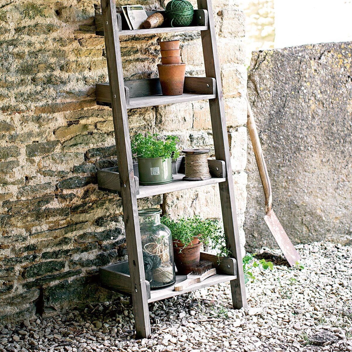 Rustikales Pflanzregal »Aldsworth« aus Holz, gefüllt mit verschiedenen Blumentöpfen und Pflanzen, steht vor einer Steinwand auf einem kiesigen Untergrund im Außenbereich.