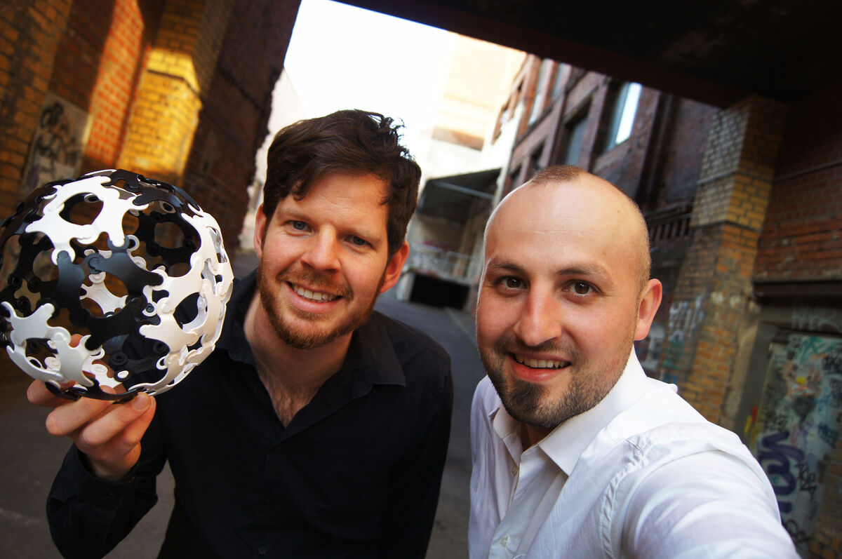 Zwei Männer posieren in einer städtischen Umgebung. Einer hält einen strukturierten Ball aus weißen und schwarzen Teilen. Sie lächeln in die Kamera.