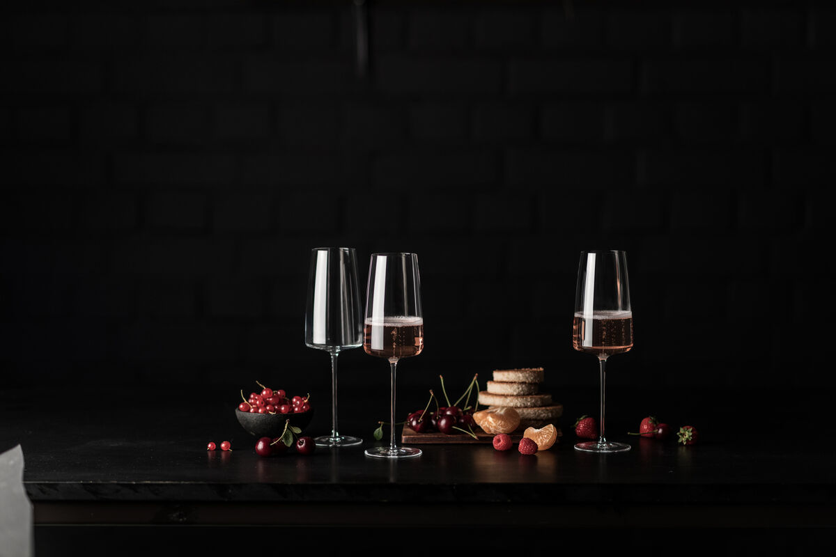 Zwei schlichte Sektgläser gefüllt mit roséfarbenem Getränk auf einem dunklen Tisch. Begleitet von ein paar frischen Kirschen und knusprigem Brot im Hintergrund. Dunkler Hintergrund, der die Gläser und die Früchte hervorhebt.