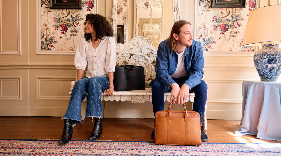 Zwei Personen sitzen auf einer weißen Bank vor einer Wand mit floralem Muster, eine hält eine schwarze Handtasche, die andere eine braune Ledertasche