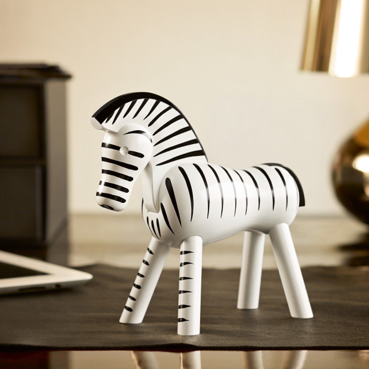 Holzfigur Zebra von Kay Bojesen, eine weiße und schwarze gestreifte Zebra-Skulptur aus Holz, die auf einer glatten Oberfläche steht. Im Hintergrund befinden sich unscharfe Elemente, darunter Möbel und eine Lampe.