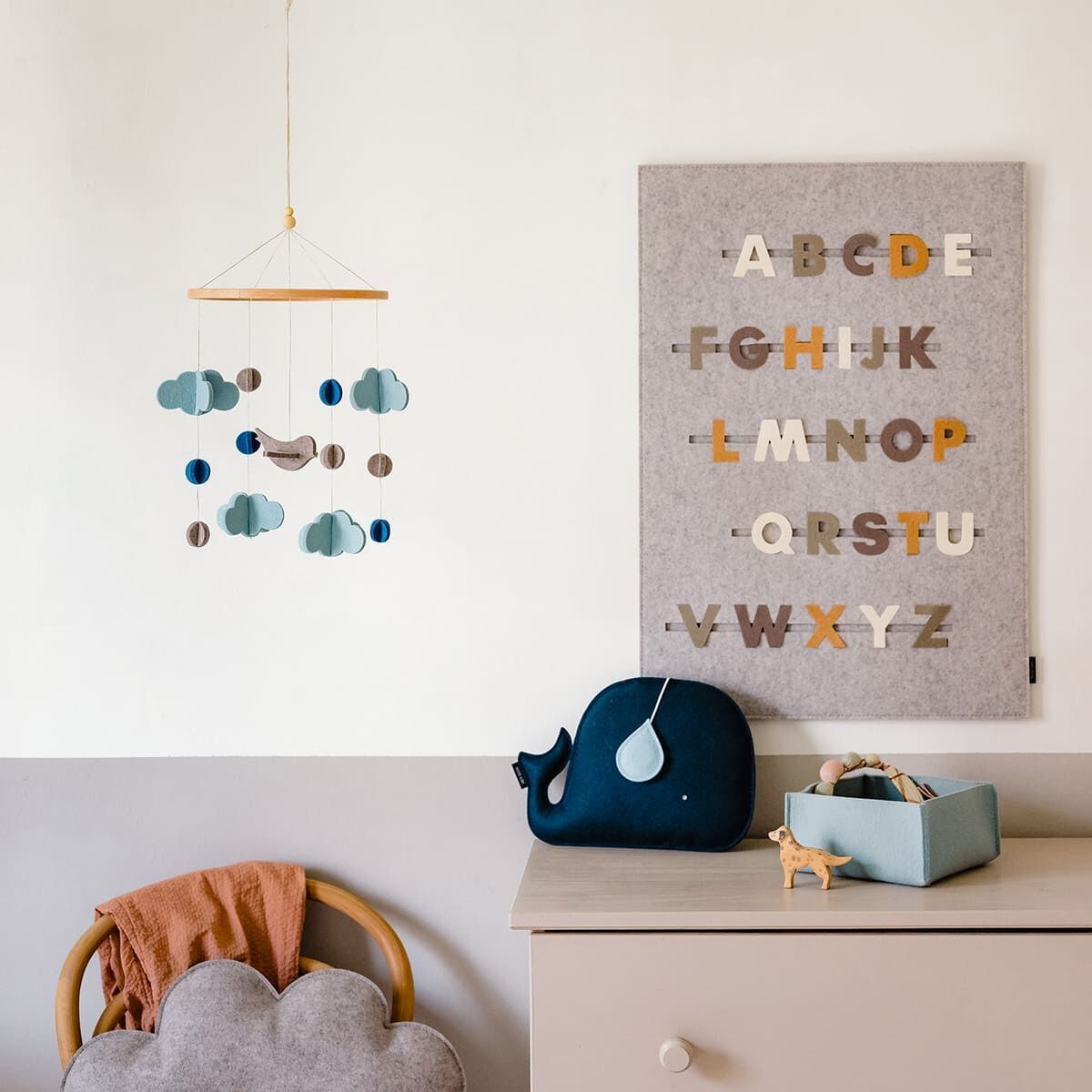 Ein hübsch eingerichtetes Kinderzimmer mit einer Filzmobilie an einem Holzrahmen, das aus grauen Wolken und blauen Tropfen besteht. Im Hintergrund hängt ein Stoffbrett mit bunten Buchstaben, die das Alphabet darstellen. Auf der Kommode stehen dekorative Elemente wie ein Spielzeugwal, ein Holzpferdchen und eine blaue Kiste.