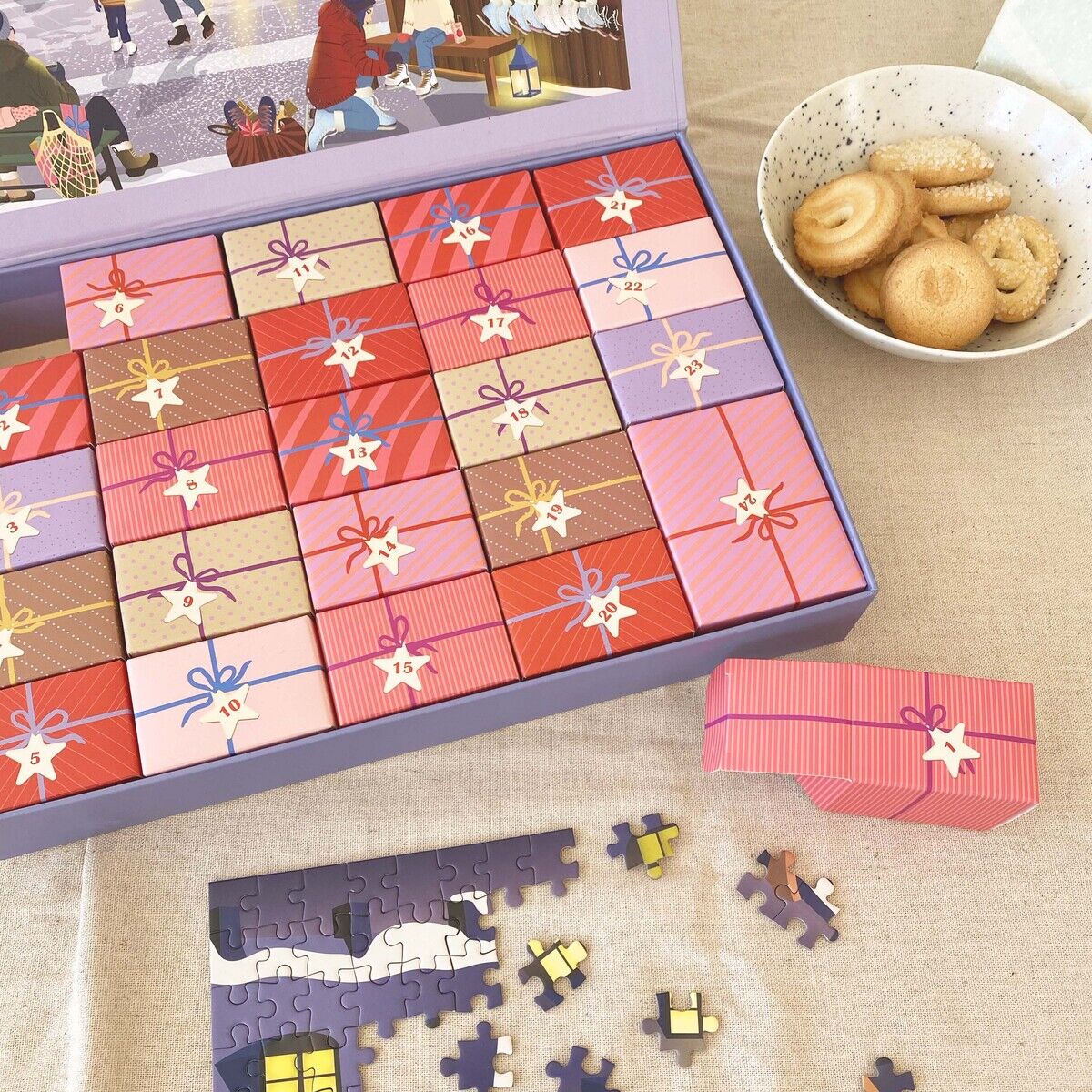Zu sehen ist der Puzzle-Adventskalender »Skate & Coffee« von ViSSEVASSE mit 24 kleinen, bunt gemusterten Schachteln in einer violetten Box. Daneben liegen Puzzle-Teile und daneben eine Schale mit hellen Keksen. Im Deckel ist eine winterliche Markt-Szene abgebildet.