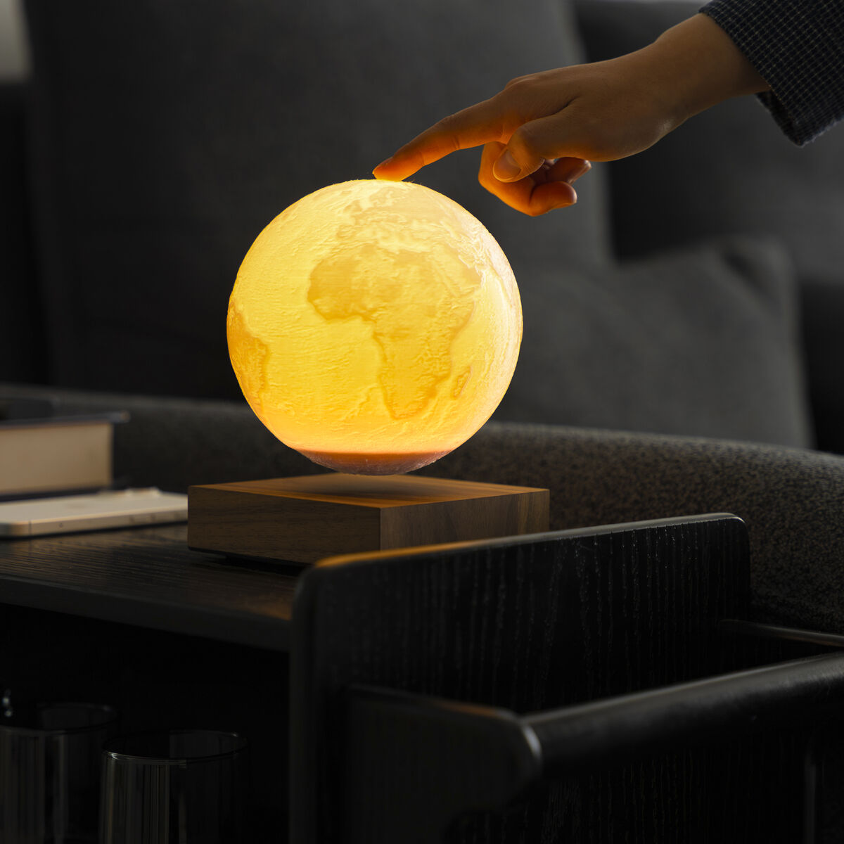 Die Szene zeigt die »Smart Earth Lamp« von Gingko Design Ltd: Eine leuchtende, gelb-orange Weltkugel-Schreibtischlampe schwebt auf einem braunen Holzsockel. Eine Hand nähert sich der Lampe, die auf einem dunklen Tisch vor einem grauen Sofa steht. Modernes, gemütliches Ambiente.