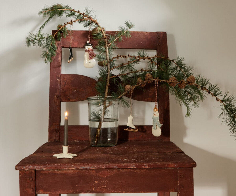 Ein rustikaler Holzstuhl steht vor einer weißen Wand. Ein Kieferzweig mit mehreren Weihnachtsanhängern aus dem Set »Winter Wunderland« befindet sich in einem Glasbehälter auf dem Stuhl. Neben dem Glas steht eine graue Kerze in einem neutralen Kerzenhalter. Die Szene strahlt eine gemütliche und festliche Atmosphäre aus.