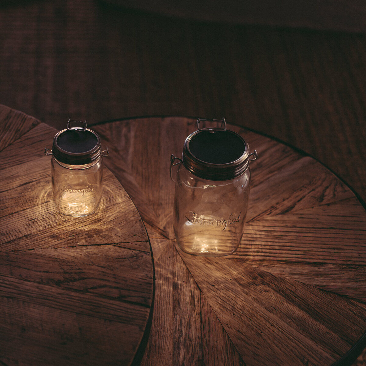 Zwei Sonnenglas-Glasbehälter mit integrierten LED-Solarlichtern, die auf einem hölzernen Tisch in einem dunklen, gemütlichen Raum stehen. Die Lichter erzeugen eine warme und stimmungsvolle Atmosphäre.