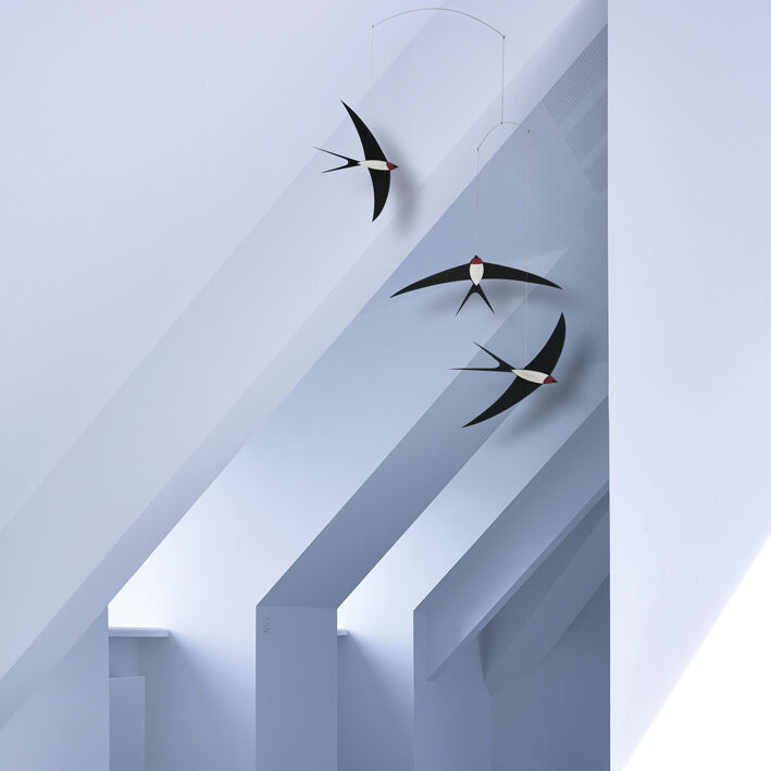 Ein Mobile mit drei stilisierten Schwalben hängt an einer Decke. Die Schwalben sind schwarz mit weißen und roten Details. Der Hintergrund ist eine weiße, minimalistische Raumdecke mit schrägen Dachflächen.