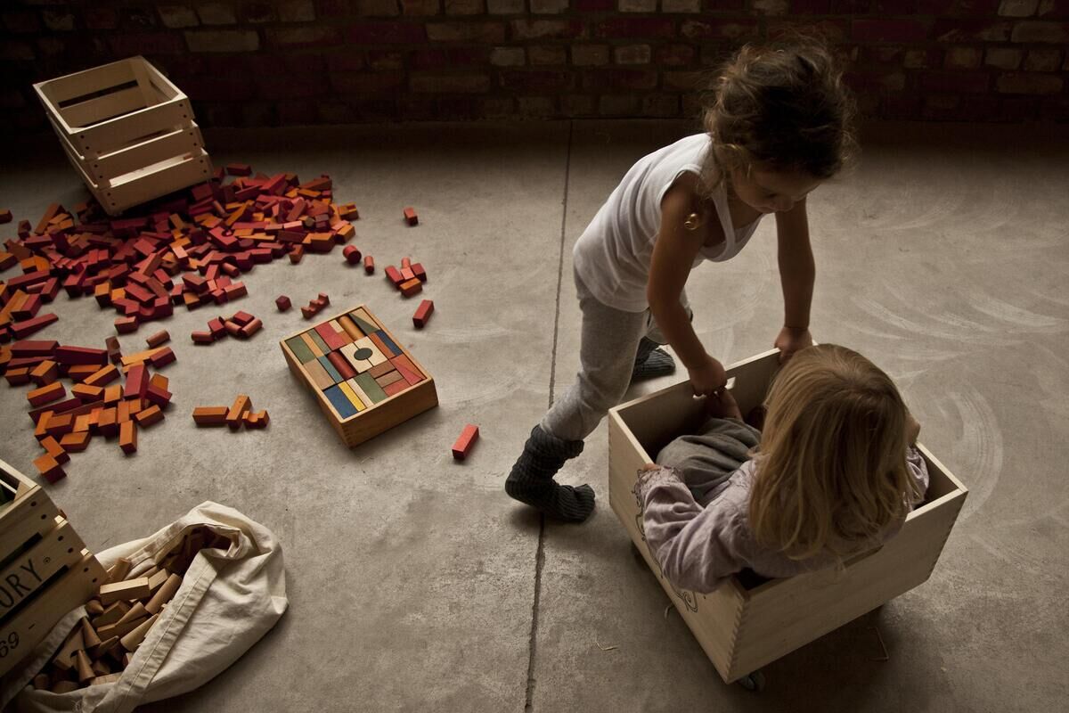 Zwei Kinder spielen mit Regenbogen Holzbausteinen. Die Bausteine sind in verschiedenen Farben und Formen. Ein Kind sitzt in einer Holzkiste, während das andere Kind die Kiste schiebt. Im Hintergrund liegen weitere Bausteine verstreut auf dem Boden.