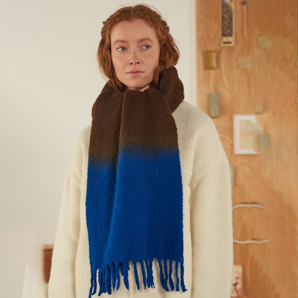 Eine Frau trägt einen dicken, handgefertigten Schal »No.698« von MOISMONT in braun und kräftigem Blau. Der Schal aus Schurwolle und Nylon, ca. 35 x 180 cm groß, hat Fransen an den Enden. Die Frau steht vor einer Holzvertäfelung, trägt einen cremefarbenen Wollpullover.
