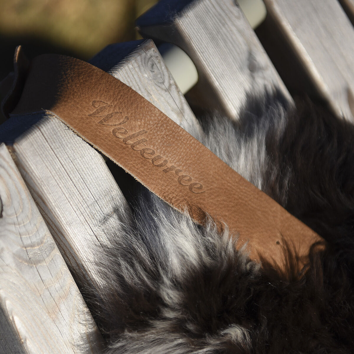 Nahaufnahme eines natürlichen Schafsfells mit einer braunen Lederschlaufe auf einer Holzbank im Freien. Die Szene wirkt gemütlich und zeigt das handwerklich gearbeitete Wohnaccessoire.