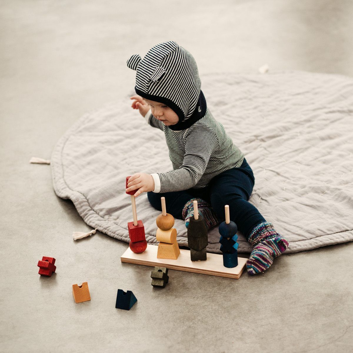 Kleinkind spielt mit einem Regenbogen Steckspielzeug aus Holz auf einer grauen Decke. Verschiedene farbige Holzfiguren sind aufgestellt und weitere liegen auf dem Boden verstreut.
