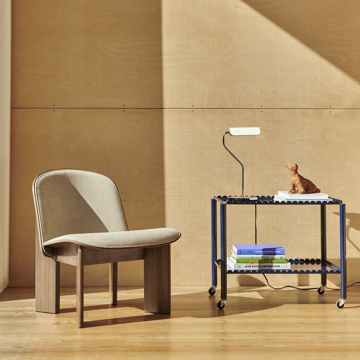 Die Apex Lampe »Table« steht auf einem blauen Metalltisch neben einem beige gepolsterten Stuhl vor einer holzverkleideten Wand. Ein Fenster mit durchsichtigen Vorhängen befindet sich auf der linken Seite des Bildes.