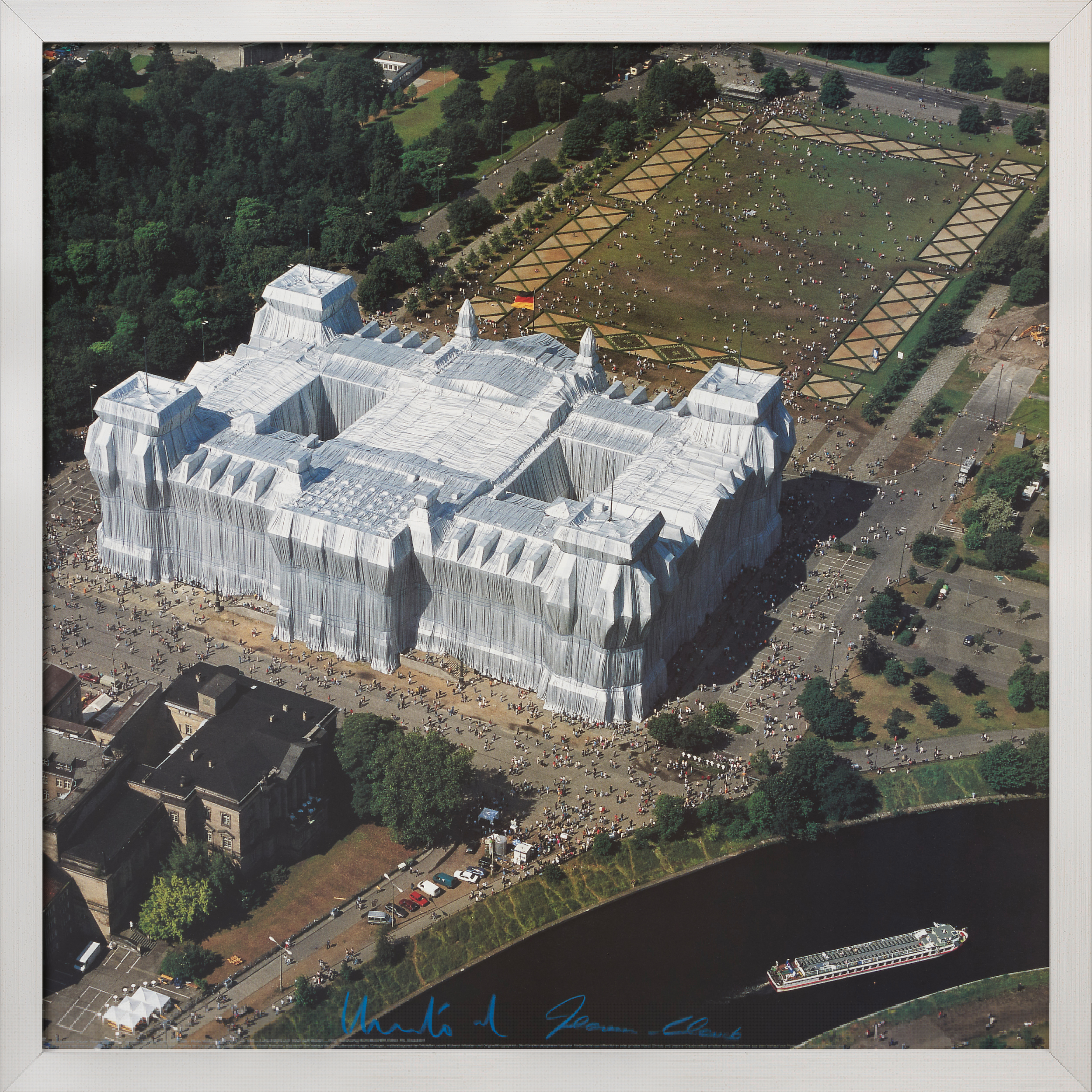 Eine gerahmte Luftaufnahme zeigt den verhüllten Berliner Reichstag von Osten nach Westen, umgeben von einem Park und einem Fluss. Die Installation ist mit weißen Stoffbahnen verkleidet und von Besuchern umgeben.