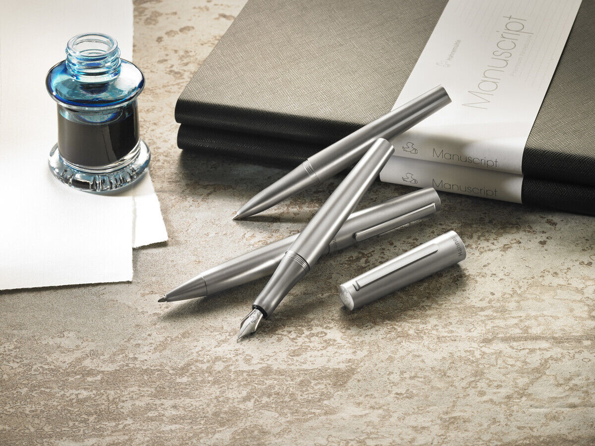 Ein Rollerball-Stift der »Bold Edition« liegt neben einem Tintenfass sowie Notizbüchern auf einem Schreibtisch. Das Bild zeigt eine elegante und hochwertige Schreibutensilie in silberner Farbe, umgeben von schwarzen und grauen Notizbüchern und Papier.