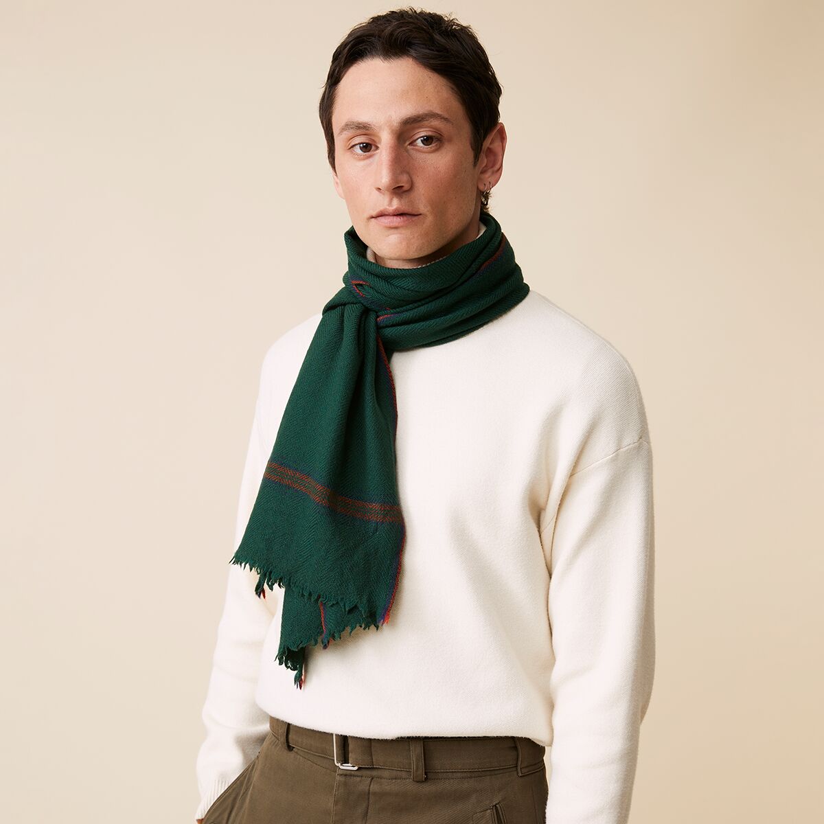 Eine Person trägt einen dunkelgrünen Schal aus 100% Schurwolle von MOISMONT um den Hals. Der Schal hat dezente Zierstreifen und ausgefranste Enden. Dazu ein cremefarbener Pullover und eine olivgrüne Hose, schlicht und stilvoll kombiniert.