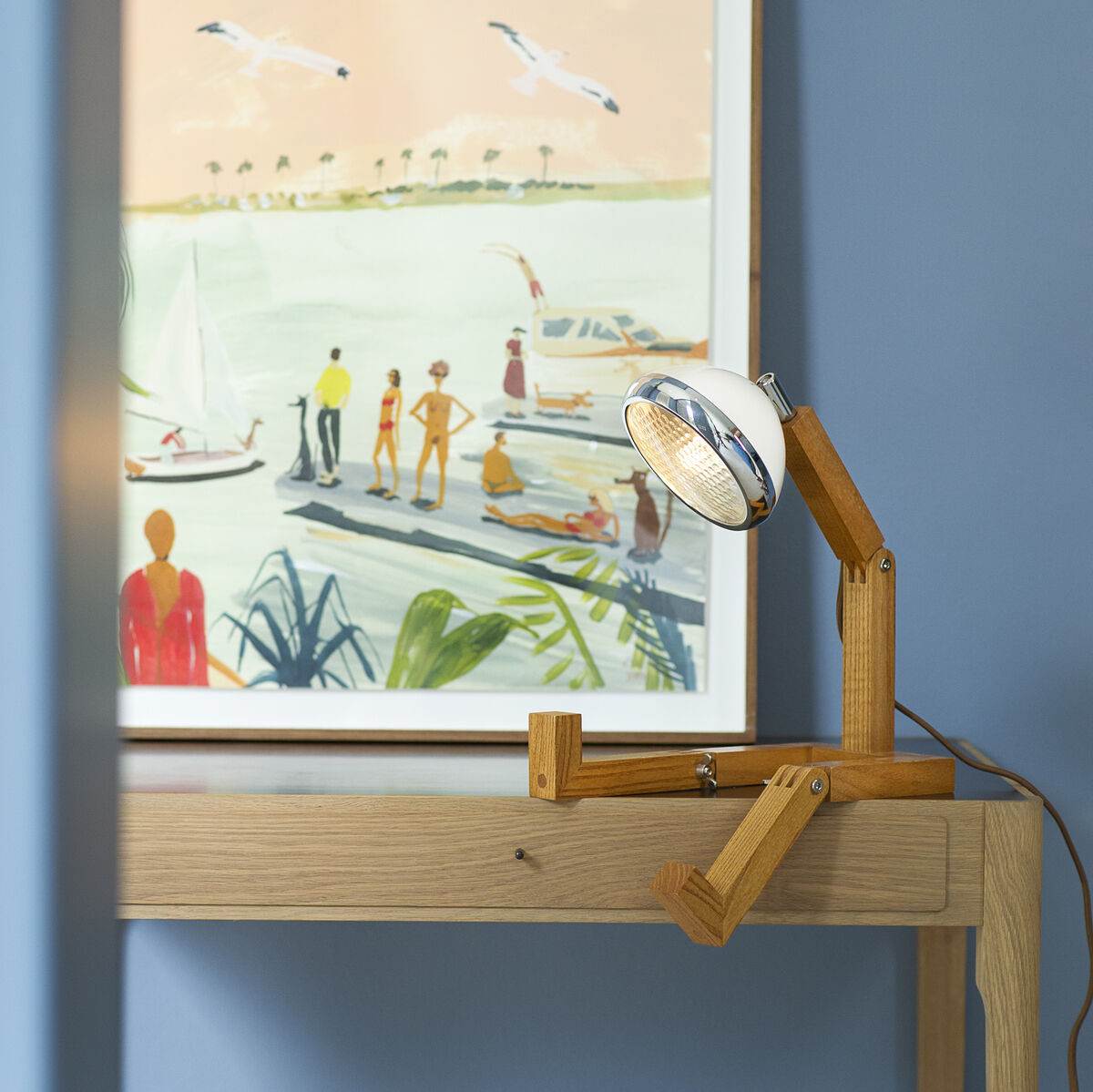 Designlampe »Mr. Wattson« aus Naturholz auf einem Holzschreibtisch vor einem Bild mit Meer- und Strandmotiven auf einer hellblauen Wand.