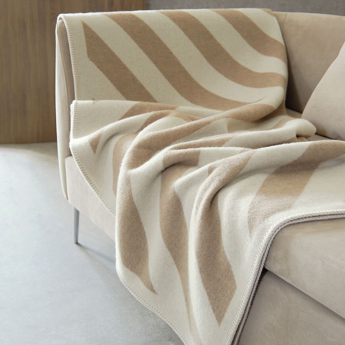 Eine beige-weiße Kaschmirdecke mit Streifenmuster liegt elegant auf einem beigen Sofa, im modernen Wohnzimmersetting.