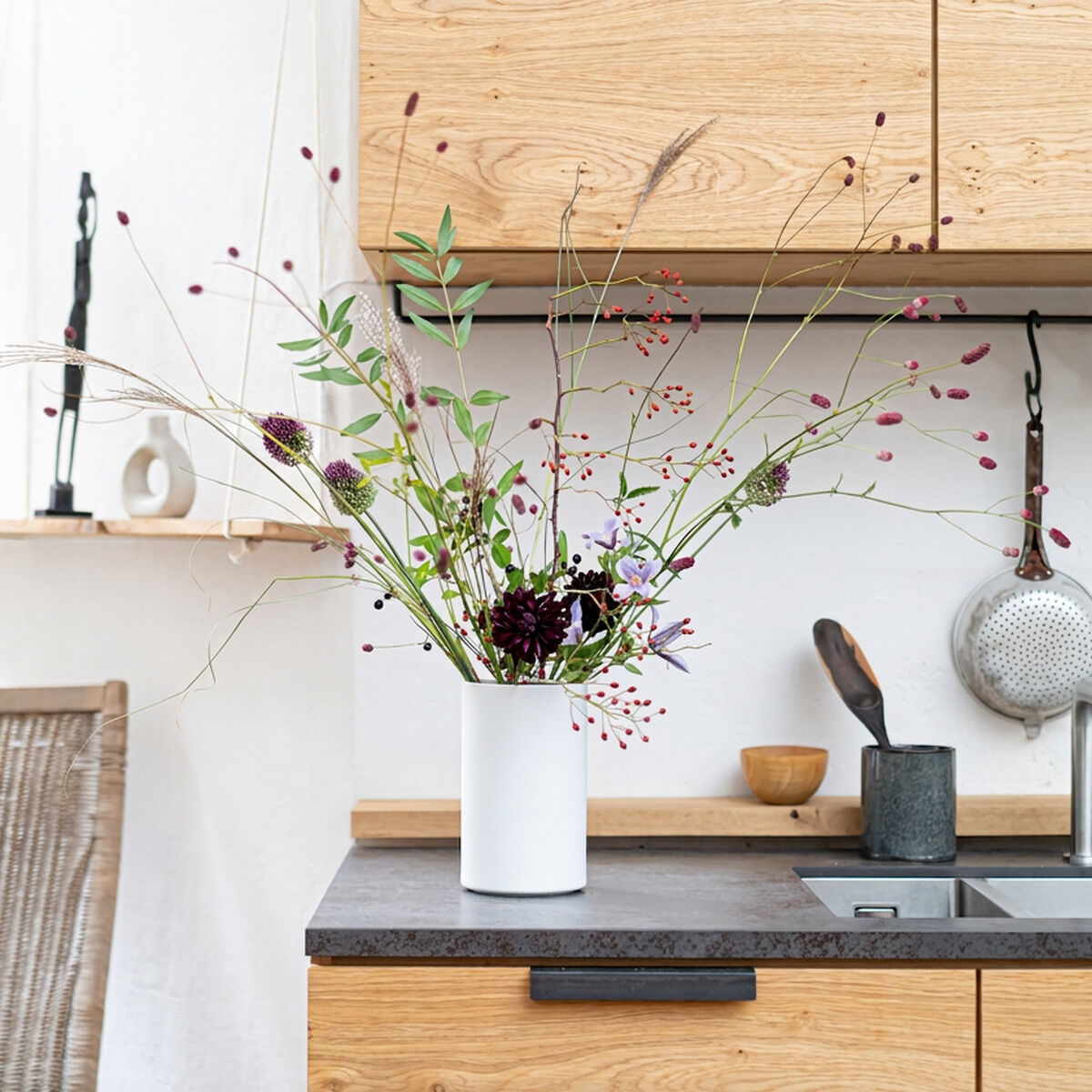 Helle, moderne Küche mit Weinkühler und Vase "KULA" von Frau Caze in weiß matt auf dunkler Arbeitsplatte. In der Vase steckt ein großes, naturinspiriertes Blumenarrangement. Holzfronten, Spülbecken und Küchenutensilien im Hintergrund.