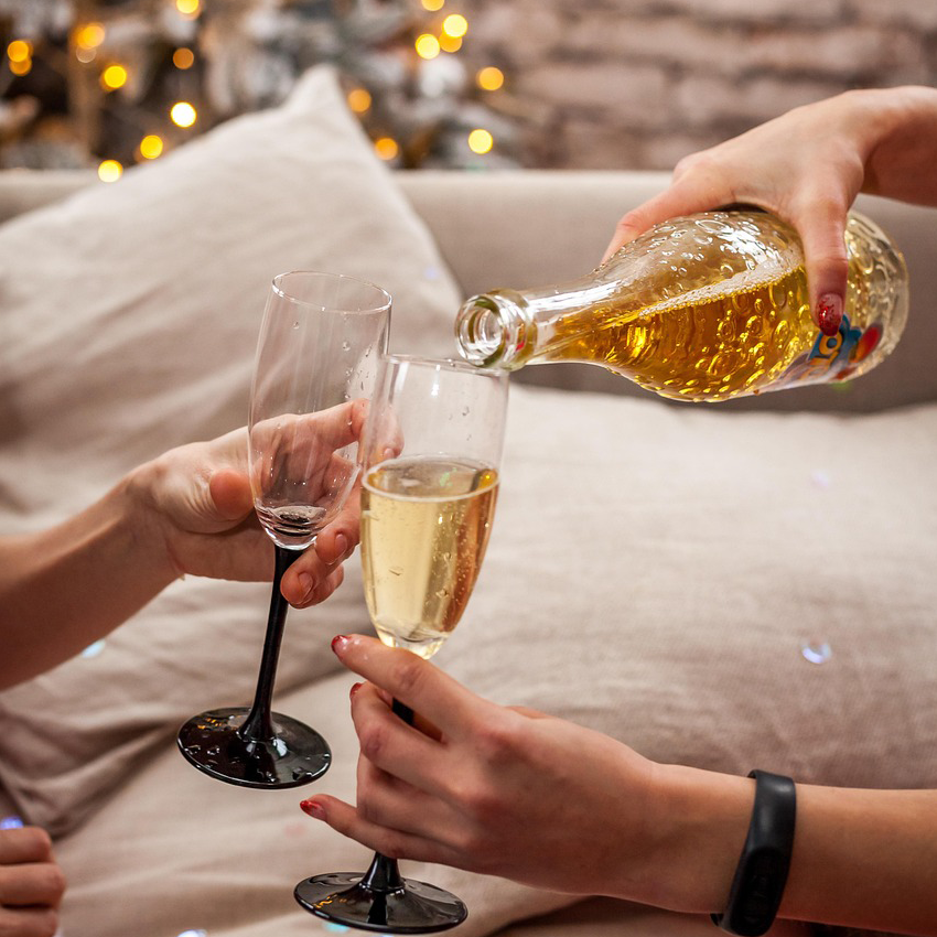 Zwei Hände halten Sektgläser, eine dritte gießt Winzerwelt Winzer Crémant in ein Glas auf einem Sofa