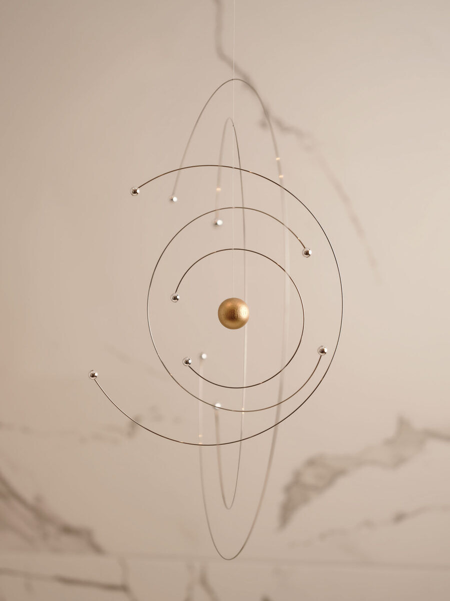 Ein modernes Mobile namens »Niels Bohr«, das physikalische Modell mit drei kreisförmigen Drähten darstellt, die sich um eine goldene Kugel im Zentrum drehen. Es ist in einem minimalistischen Stil gehalten und an einer unsichtbaren Schnur aufgehängt. Der Hintergrund ist ein einfacher beigefarbener Raum mit subtilen Marmorierungen.