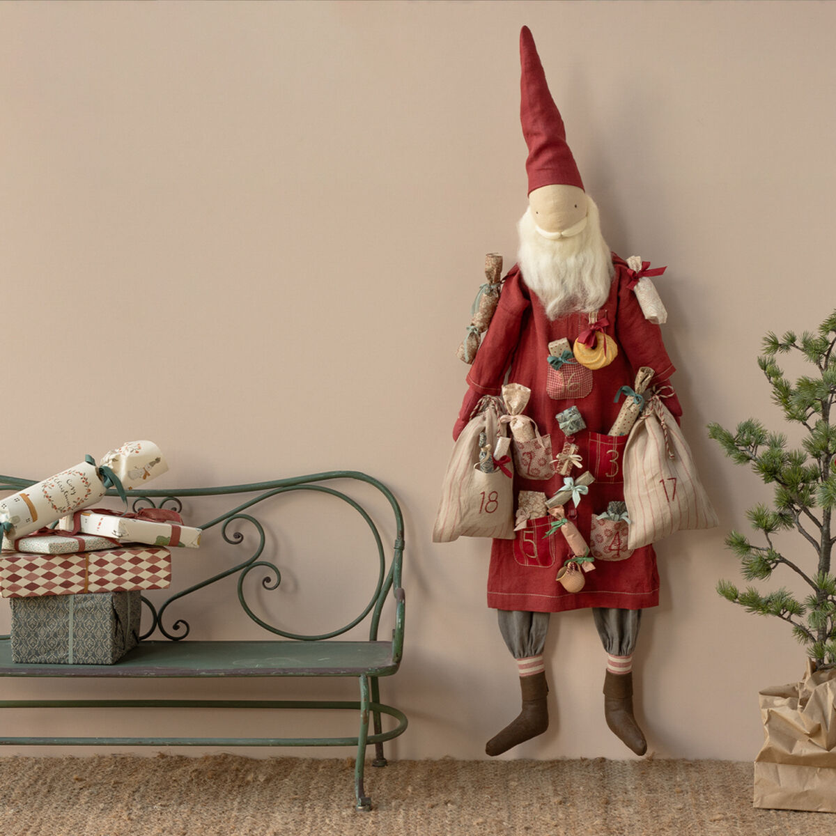 Ein adventskalender in Form eines freundlichen Weihnachtsmanns aus Stoff hängt an einer beigefarbenen Wand. Der Santa trägt einen roten Mantel und eine Mütze, an ihm sind nummerierte Beutel und kleine Geschenke befestigt. Daneben steht eine dekorative Metallbank mit eingepackten Geschenken und ein kleiner Tannenzweig. Die Szene verbreitet gemütliche Weihnachtsstimmung.