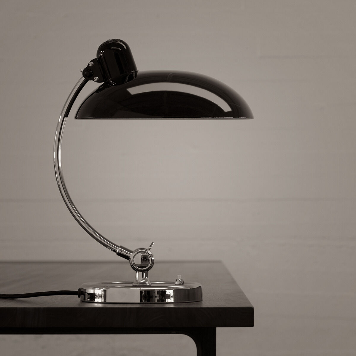 Schwarze Tischlampe »Kaiser Idell« mit silbernem Fuß und gewölbtem Schirm, auf einem minimalistischen Holztisch platziert. Schlichte Hintergrundfarbe in Grau.