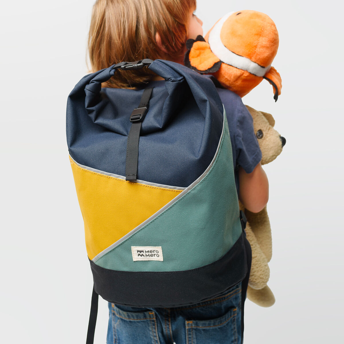 Ein Kind trägt einen Rucksack mit Farbblockdesign in Blau, Gelb und Türkis. Es hält ein orange-weißes Stofftier, das wie eine Giraffe aussieht, sowie ein braunes Stoffbärchen.