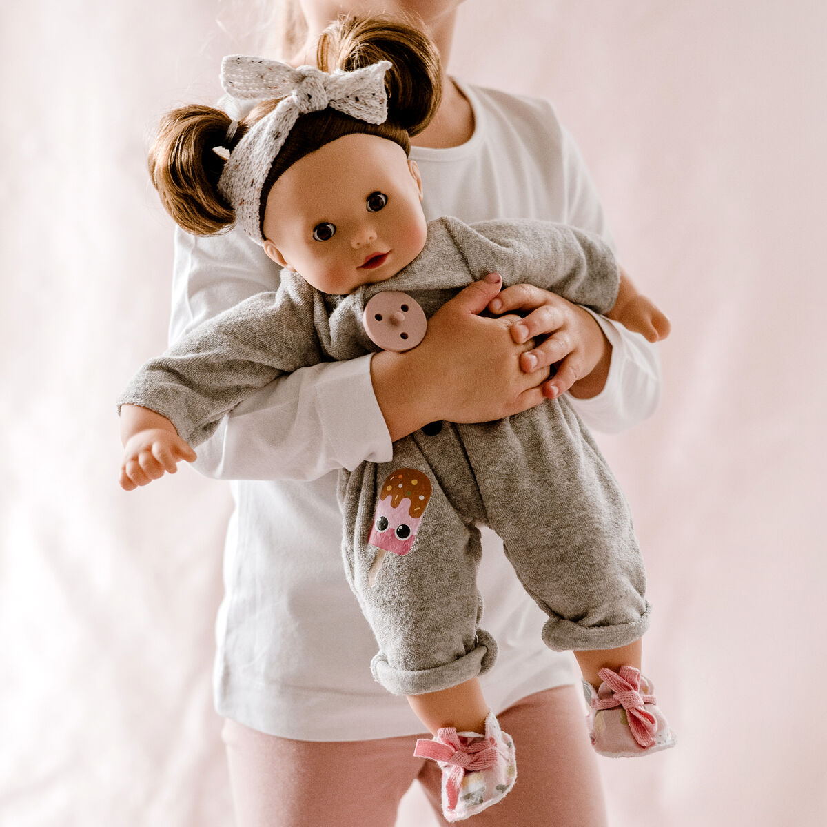 Ein Kind hält die Stoffpuppe »Maxy Muffin Popsicle« im Arm. Die Puppe trägt einen grauen Strampler mit Eiscreme-Motiv, rosa Hausschuhe und ein Haarband. Im Hintergrund ist ein neutraler, heller Hintergrund zu sehen.