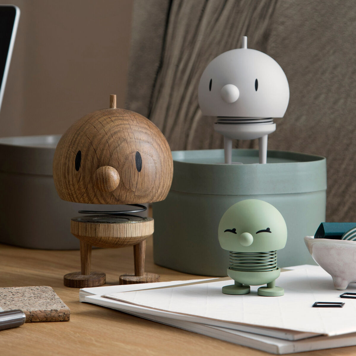 Ein Bild zeigt drei Hoptimist Bumble Figuren in unterschiedlichen Farben (Braun, Weiß, Grün) auf einem Schreibtisch. Im Hintergrund sind Schreibtischartikel und ein Laptop zu sehen.