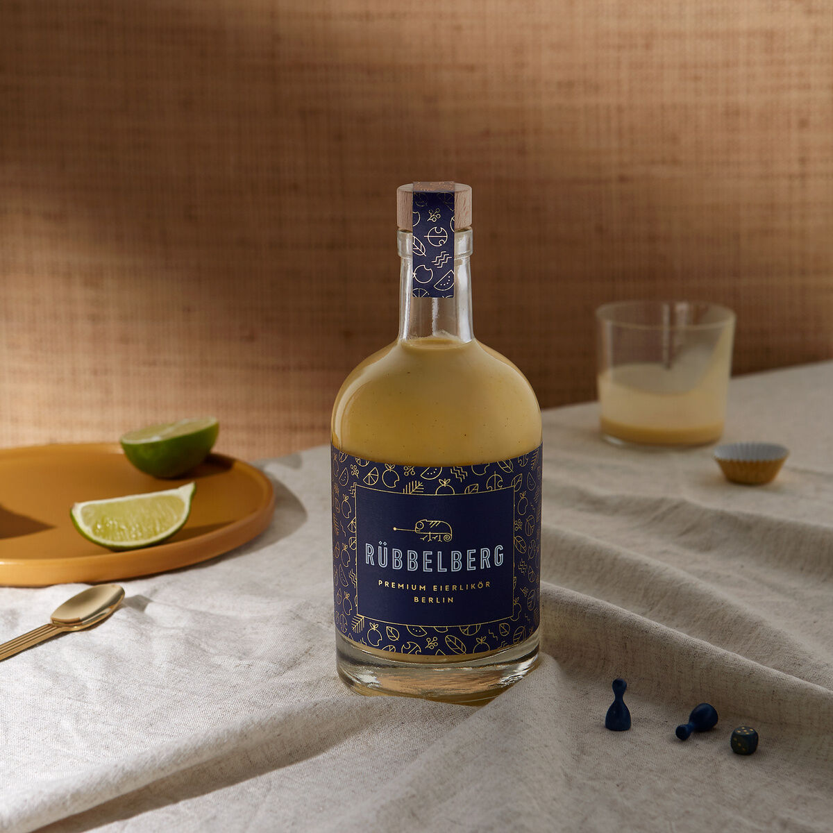 Eine Flasche Premium Eierlikör von Rübbelberg steht auf einem Tisch mit einem Glas und Limettenscheiben. Die Flasche hat ein edles dunkelblaues Etikett mit goldenen Details, und im Hintergrund ist ein Stoff mit natürlichen Farben zu sehen.